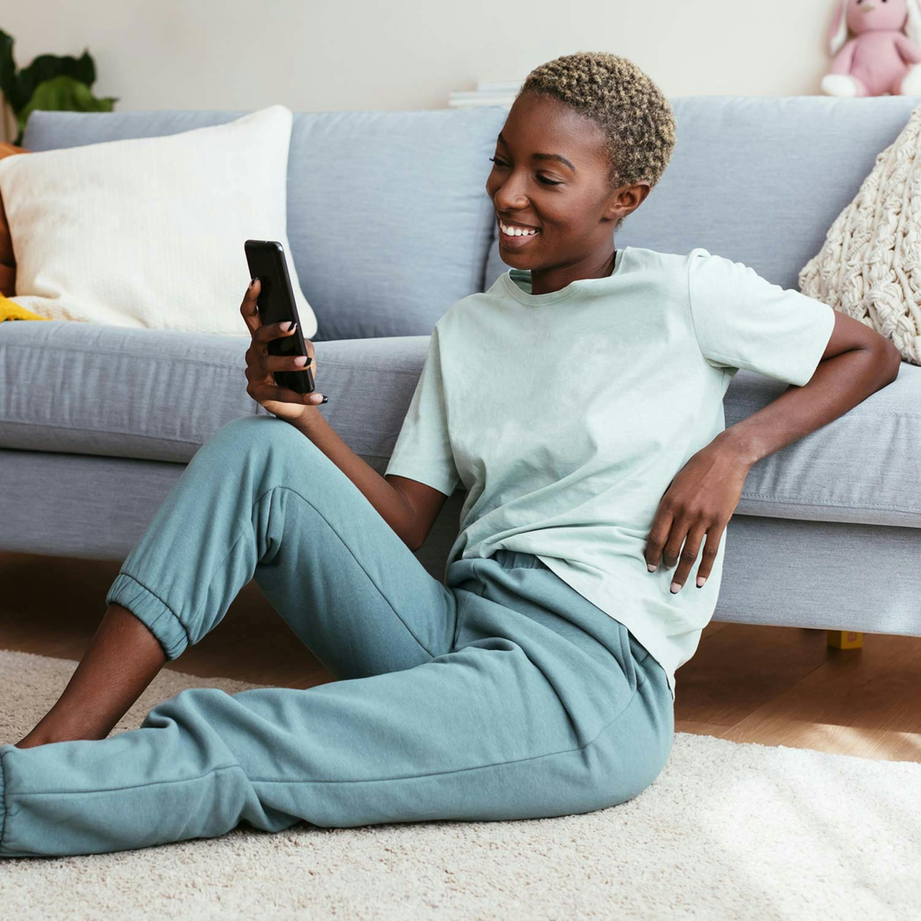 Lächelnde Frau mit Smartphone vor Couch am Boden sitzend 
